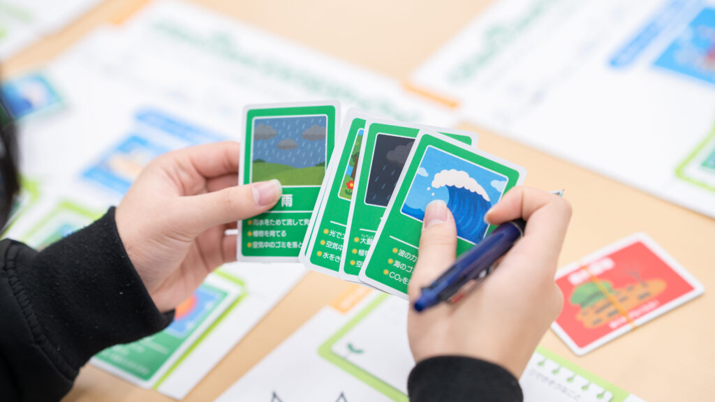 サステナブルなまちづくり教材「サスネタ！」で、雨や波などの条件カードを手にワークシートへ書き出す場面