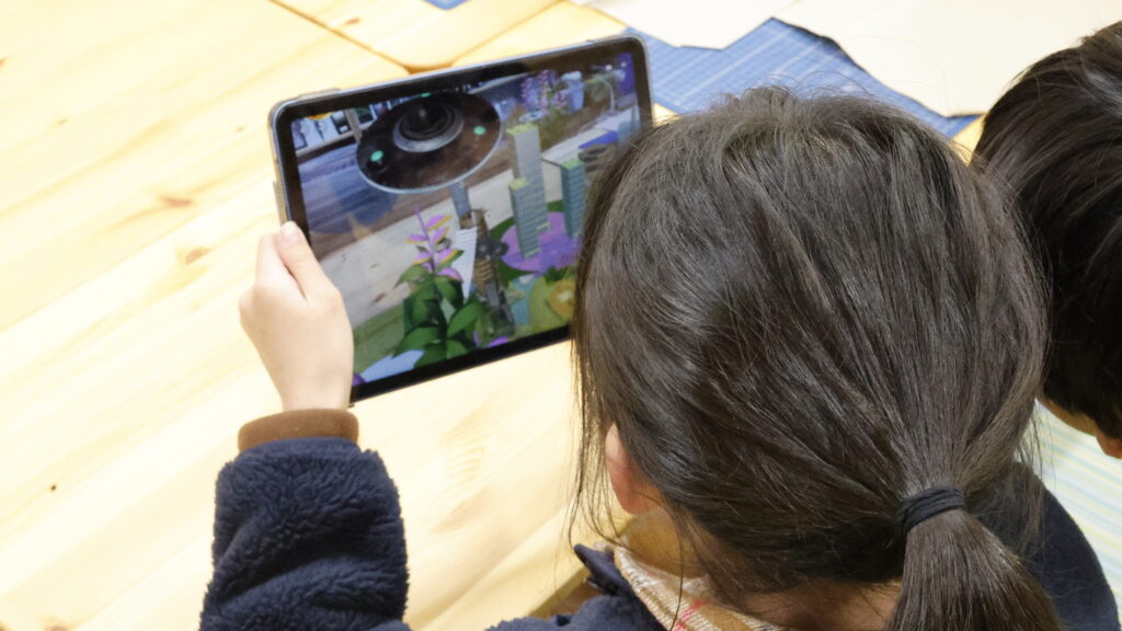 サステナブルなまちづくり教材「サスネタ！」で作った都市の3Dモデルを、タブレットのARで投影して配置やデザインを検討している場面
