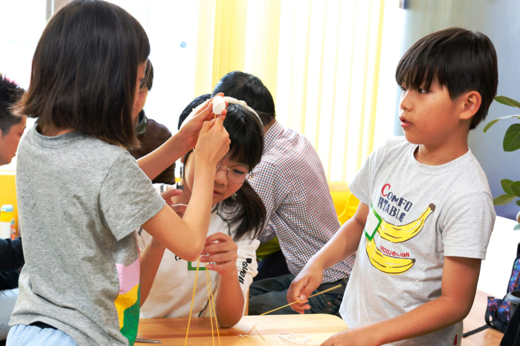 小学生たちがチームで協力し、パスタとマシュマロを使ってタワーを組み立てるSTEAM教育ワークショップの様子。