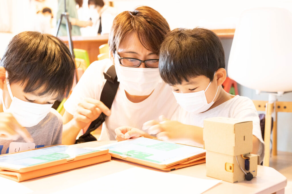 親子がロボットプログラミング教室でタブレットを使って学ぶ様子。小学生がプログラムを組み立てるSTEAM教育の現場。