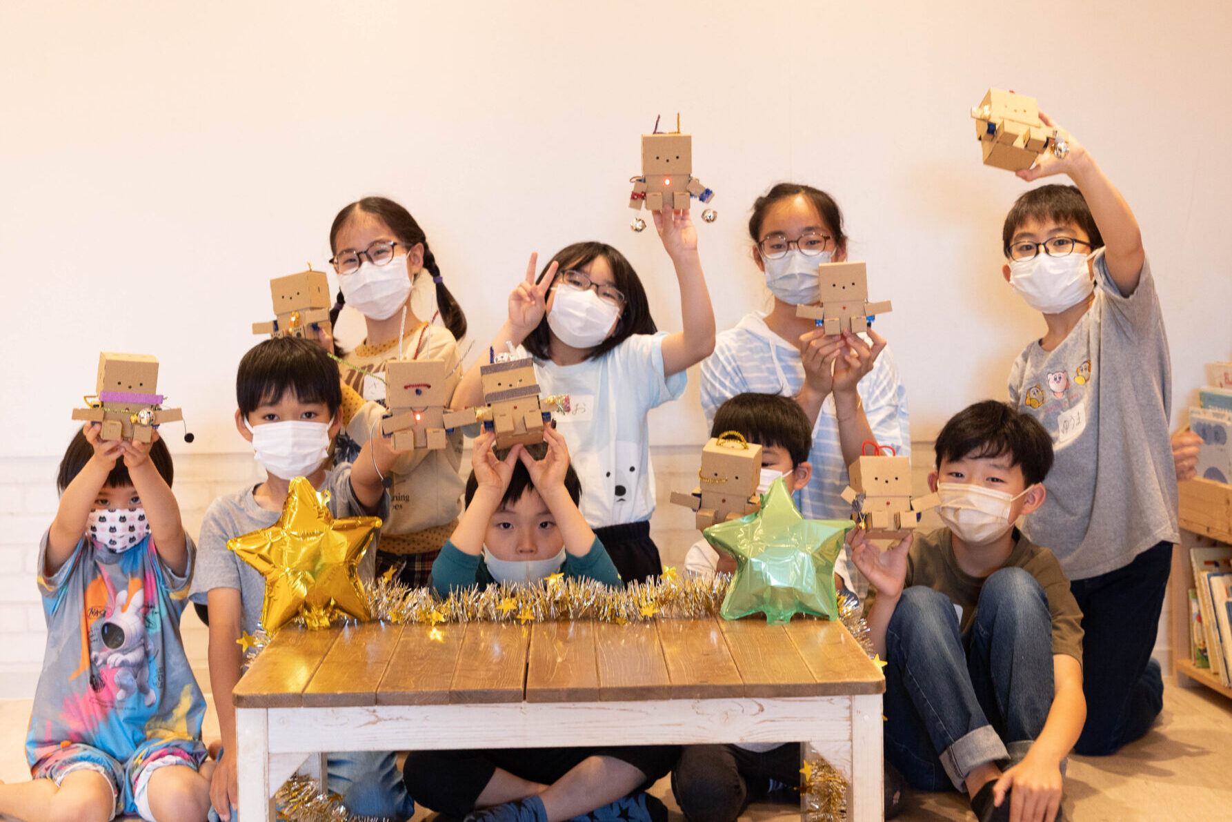 小学生たちが自作の段ボール製ロボットを持ち寄り、STEAM工作教室の活動成果を笑顔で発表する集合写真。