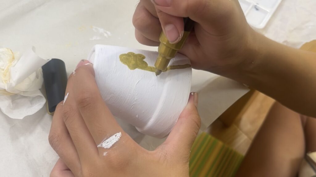 白く塗った植木鉢に金色の模様を描く子どもの手元。風鈴づくりの自由なデザイン工程