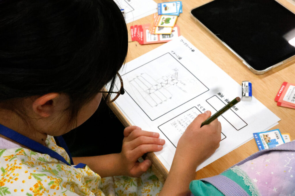 建物のアイデアスケッチと特徴をワークシートにまとめる小学生。自由研究で未来のまちをデザイン。