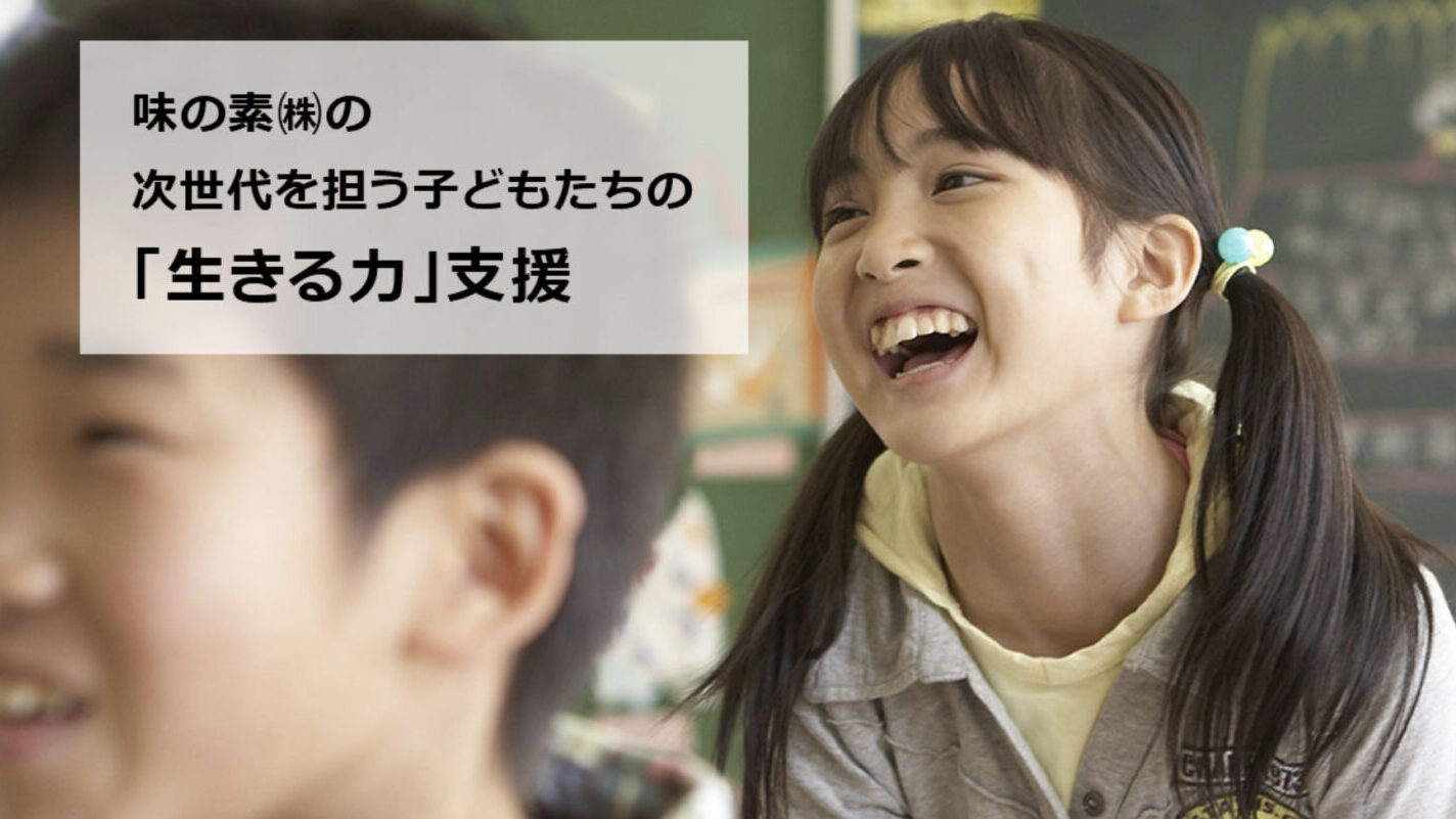 味の素の次世代支援活動を象徴するビジュアル。教室で笑顔を見せる女の子と、手前に映る子どもたちの姿。「生きる力」支援を掲げたメッセージが添えられている。